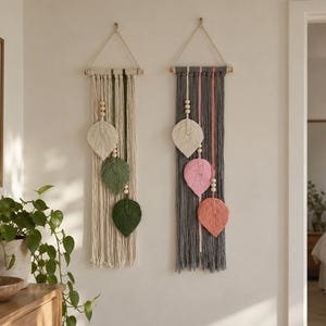 May include: Two macrame wall hangings suspended from wooden dowels with rope. The left piece features cream and green cord with three leaf-shaped accents and wooden beads. The right piece features gray, pink, and coral cord with similar leaf accents.