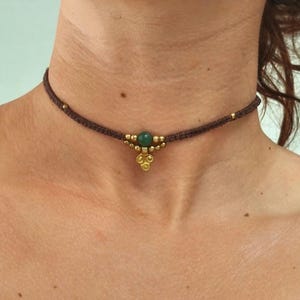 May include: A brown choker necklace with a central green stone and gold-coloured accents. The necklace is made of woven material and features small gold beads along the band. The pendant has a cluster design.