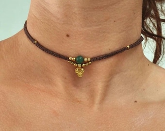 Può includere: Una collana girocollo marrone con una pietra verde centrale e dettagli color oro. La collana è realizzata in materiale intrecciato e presenta piccole perline dorate lungo la fascia. Il ciondolo ha un design a grappolo.