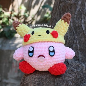 May include: A handmade pink crochet Kirby plush wearing a yellow Pikachu hat with red accents. The plush has black eyes, a small mouth, and red feet. The text "pandie.crochet" is visible.