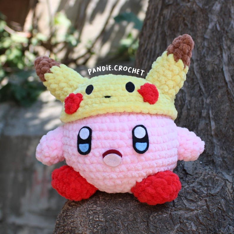 May include: A handmade pink crochet Kirby plush wearing a yellow Pikachu hat with red accents. The plush has black eyes, a small mouth, and red feet. The text "pandie.crochet" is visible.