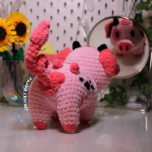 May include: A pink crocheted pig with black eyes and a curly tail. The pig is standing on a white surface and looking at its reflection in a mirror.