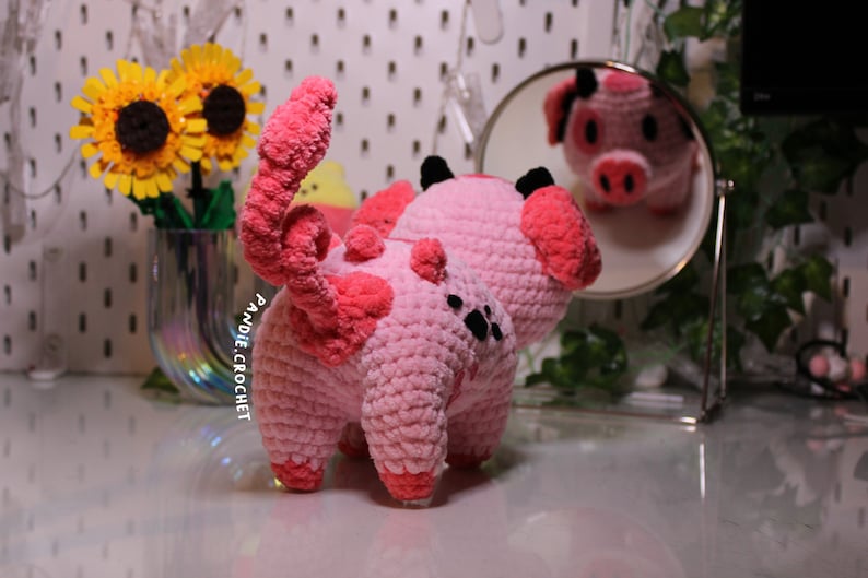 May include: A pink crocheted pig with black eyes and a curly tail. The pig is standing on a white surface and looking at its reflection in a mirror.