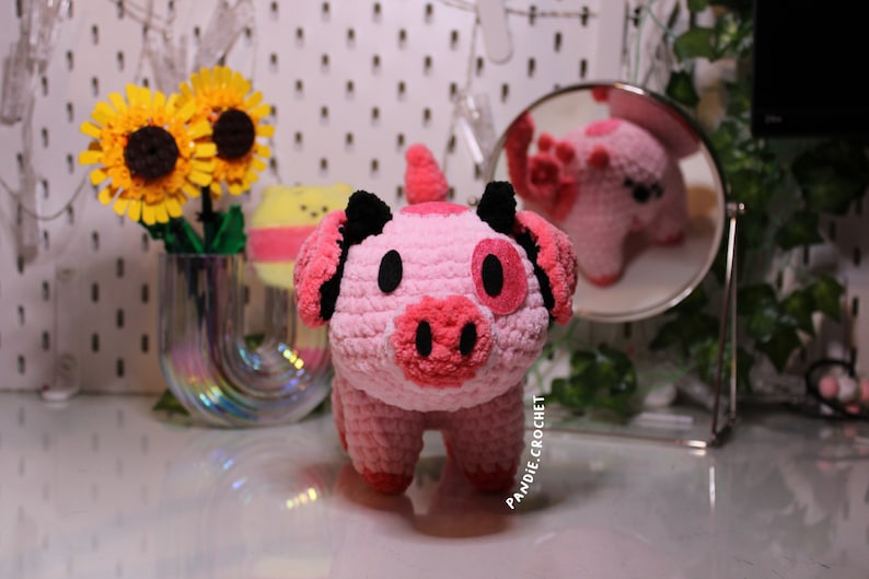 May include: A pink crocheted pig with black ears and a pink snout. The pig is standing on a white surface with a sunflower in the background. The text "Pandle Crochet" is visible on the pig's leg.
