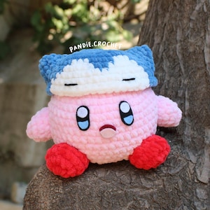 May include: A handmade pink crochet character with blue eyes, small arms, and red feet. The character is wearing a blue and white hat. The text "pandie.crochet" is visible in the image.