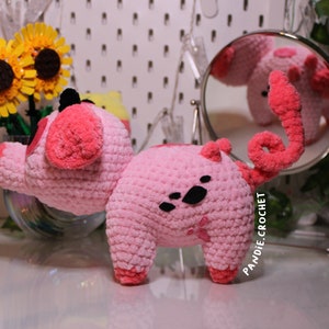 May include: Pink crocheted pig plush toy with black eyes and a curly tail. The pig is standing on a white surface with a mirror reflecting the pig in the background.