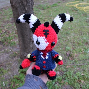May include: A handmade crochet doll with a distinctive design. The doll has a red and white head with black and white striped ears. It is dressed in a blue jacket with red details and black shoes. The doll is held against a blurred green backdrop.