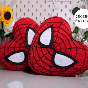 May include: Two red crocheted heart-shaped cushions with black webbing and white felt eyes. The cushions are on a white surface with a flower arrangement in the background.