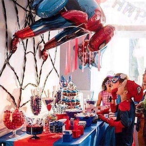 Puede incluir: Un globo de Spiderman rojo y azul cuelga de una decoraci&oacute;n de telara&ntilde;a negra sobre una mesa de postres con dulces rojos y azules, cupcakes y otras golosinas. Ni&ntilde;os vestidos de Spiderman se re&uacute;nen alrededor de la mesa.