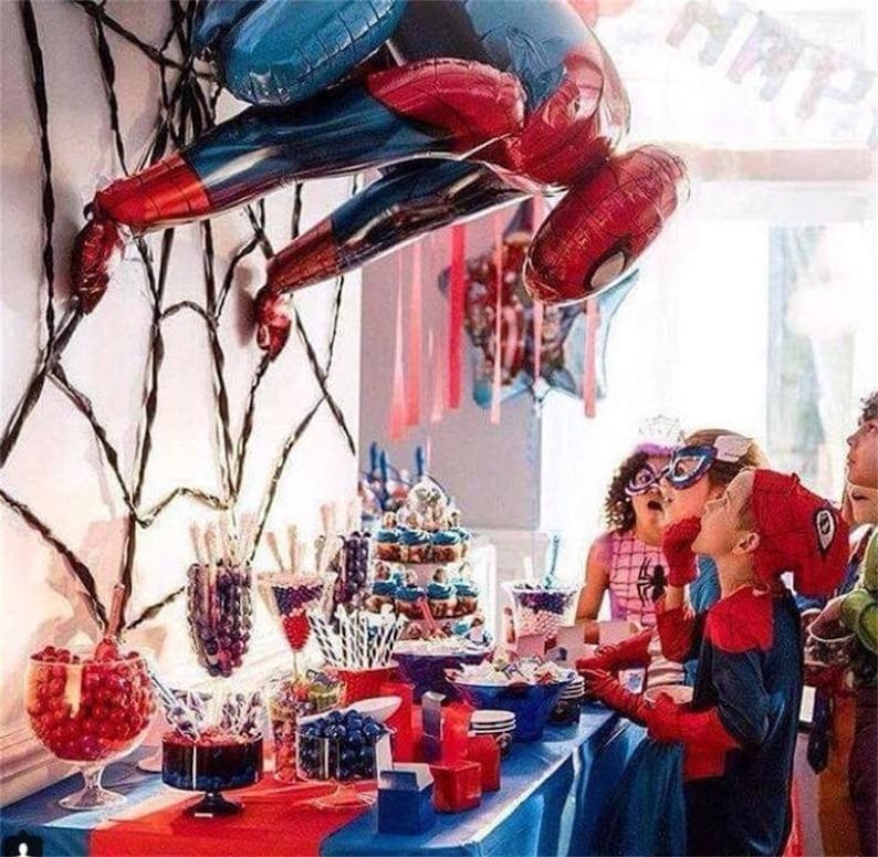 Puede incluir: Un globo de Spiderman rojo y azul cuelga de una decoraci&oacute;n de telara&ntilde;a negra sobre una mesa de postres con dulces rojos y azules, cupcakes y otras golosinas. Ni&ntilde;os vestidos de Spiderman se re&uacute;nen alrededor de la mesa.