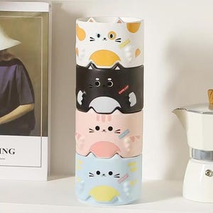 May include: A stack of four ceramic mugs, each featuring a different cat face design in white, black, pink, and light blue. Each mug has a small rectangular label with the text CUTE CAT printed on the side, and raised paw-like details.