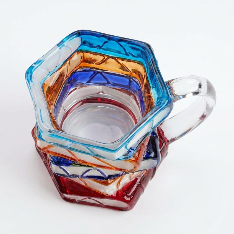 May include: A colorful, stacked glass pitcher with a clear handle. The pitcher features a hexagonal design with layers of blue, orange, red, and clear glass. The pitcher is suitable for serving beverages.