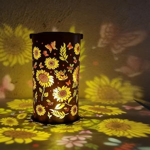Pode incluir: Uma lanterna cilíndrica castanho-escura com um design de girassóis e borboletas. A lanterna emite uma luz amarela quente, projetando padrões de girassóis e borboletas na superfície circundante. O design inclui recortes detalhados de girassóis, folhas e borboletas.