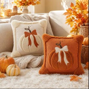 May include: Two square throw cushions featuring a raised, textured pumpkin design. One cushion is cream-coloured with an orange pumpkin and bow, while the other is orange with a cream-coloured pumpkin and bow. Both have fuzzy corner accents.