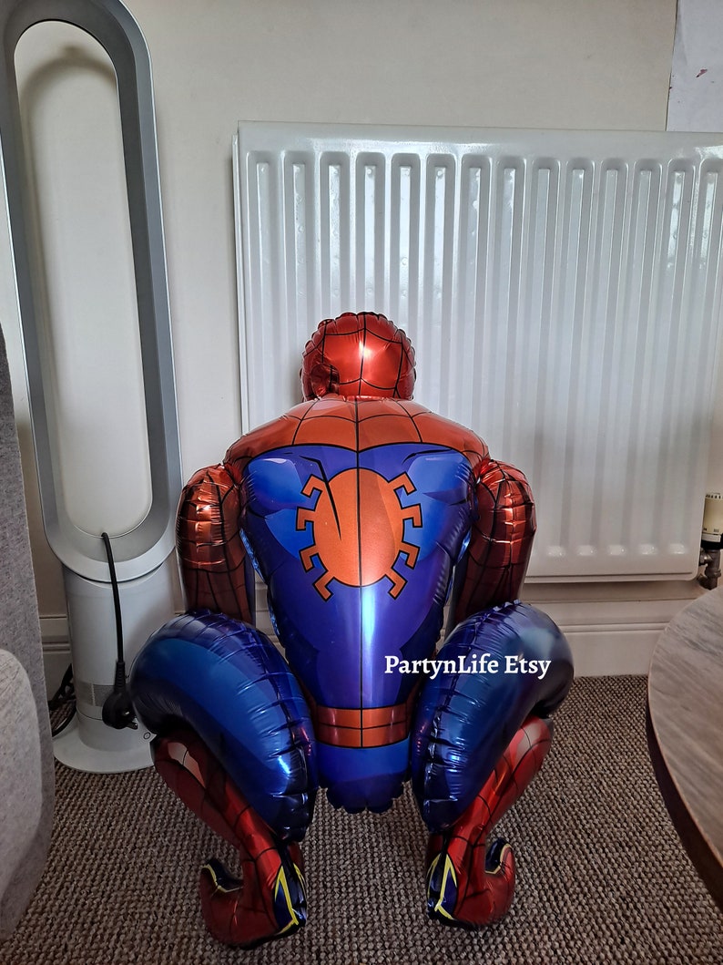 Puede incluir: Un globo de Spider-Man rojo y azul con un dise&ntilde;o de telara&ntilde;a blanca. El globo est&aacute; en posici&oacute;n de cuclillas con los brazos doblados y las piernas separadas. El globo est&aacute; sobre una alfombra con un radiador blanco en el fondo. El texto "PartyLife Etsy" es visible en el globo.