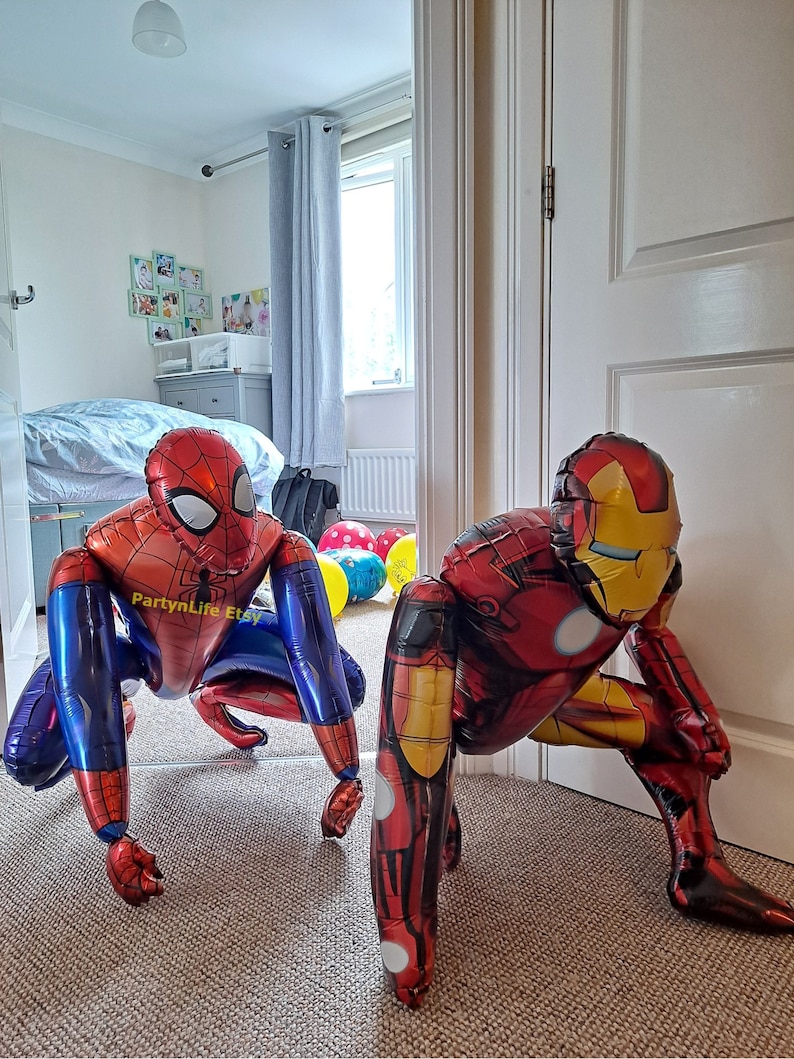 Puede incluir: Dos grandes figuras inflables de superh&eacute;roes, un Iron Man rojo y dorado y un Spiderman rojo y azul, est&aacute;n en posici&oacute;n de cuclillas en un suelo de moqueta. Las figuras est&aacute;n hechas de aluminio y tienen un acabado brillante.