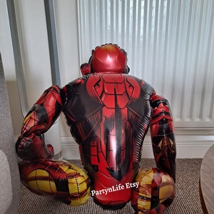 Puede incluir: Una figura inflable de Iron Man de color rojo y dorado en posici&oacute;n de cuclillas. La figura est&aacute; hecha de un material brillante y met&aacute;lico y tiene un dise&ntilde;o detallado. La figura est&aacute; sentada en una alfombra con un radiador blanco en el fondo. El texto "PartyLife Etsy" es visible en la pierna de la figura.