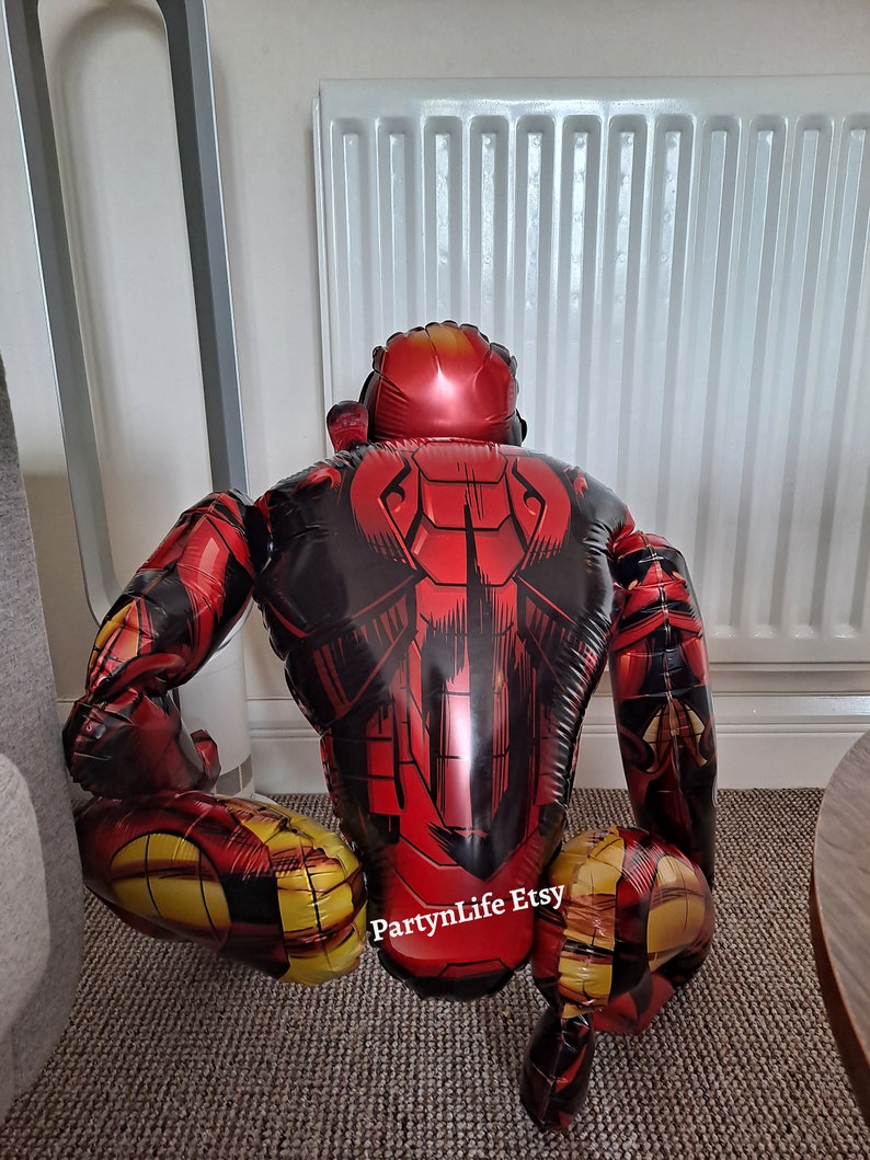 Puede incluir: Una figura inflable de Iron Man de color rojo y dorado en posici&oacute;n de cuclillas. La figura est&aacute; hecha de un material brillante y met&aacute;lico y tiene un dise&ntilde;o detallado. La figura est&aacute; sentada en una alfombra con un radiador blanco en el fondo. El texto "PartyLife Etsy" es visible en la pierna de la figura.