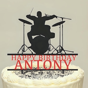 Può includere: Una torta glassata bianca con un cake topper nero e rosso. Il topper presenta la silhouette di un batterista che suona una batteria, con le parole "HAPPY BIRTHDAY ANTONY" in rosso. La torta è su un supporto nero in ferro battuto.