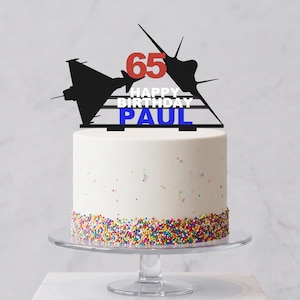 May include: A black cake topper featuring two fighter jet silhouettes, the red number 65, and the white text HAPPY BIRTHDAY above the blue text PAUL, all positioned on a white cake with colorful sprinkles on a clear glass pedestal stand.
