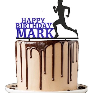 May include: A blue and black cake topper with the text "Happy Birthday Mark" and a silhouette of a runner. The topper is on top of a chocolate cake with chocolate ganache.