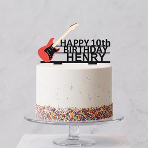 May include: A black cake topper featuring a red electric guitar graphic and the text HAPPY 10th BIRTHDAY HENRY, positioned on a white frosted cake with colourful sprinkles around the base, resting on a clear glass pedestal stand.