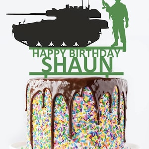 May include: Green silhouette of a tank and a soldier with a rifle, with the words "Happy Birthday Shaun" on a cake topper. The cake is covered in colorful sprinkles and has chocolate frosting.