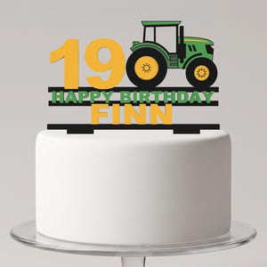 May include: A cake topper featuring a green tractor, the yellow number 19, and the text HAPPY BIRTHDAY FINN in green and yellow lettering. The design is mounted on black horizontal bars, creating a decorative accent for a birthday cake.
