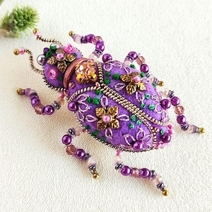 Op de afbeelding: Een handgemaakte, paarse vilten keverbroche, ingewikkeld versierd met kralen, pailletten en bloemenborduurwerk. De broche heeft goudkleurige metalen accenten en kralenpoten, wat een uniek en gedetailleerd accessoire creëert. Het ontwerp bevat roze en groene accenten.