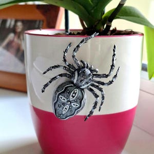 Könnte beinhalten: Ein dekoratives Spinnenornament, das an einem zweifarbigen Blumentopf befestigt ist. Der Topf ist weiß mit einem roten Band und enthält eine grüne Pflanze. Die Spinne ist silber und schwarz, mit detaillierten Beinen und Körper.