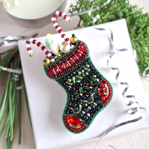 May include: A beaded Christmas stocking brooch featuring a black and green body with red accents. The top is decorated with red and white striped candy cane elements, a white pearl, and a small green bead, all crafted with intricate beadwork.