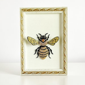Front view of a small framed art piece showcasing a 3D bead-embroidered honey bee on an off-white fabric background.