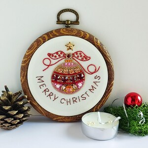 May include: A circular embroidery hoop with a wood-grain frame containing white fabric. The fabric features a beaded Christmas ornament design in shades of red, pink, and gold, with the text "MERRY CHRISTMAS" embroidered in dark brown thread.