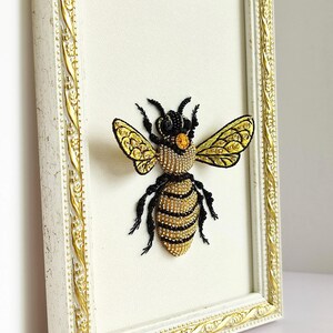 Cottagecore decor styling of a framed insect embroidery alongside a small glass vase of white flowers.