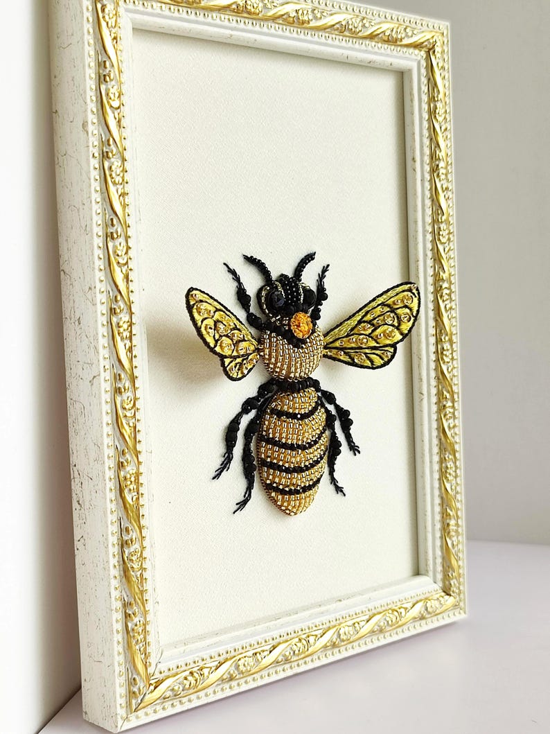 Cottagecore decor styling of a framed insect embroidery alongside a small glass vase of white flowers.