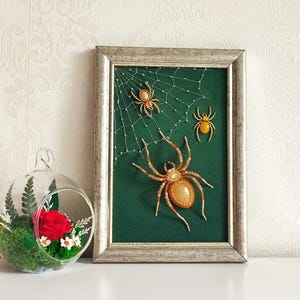 Può includere: Un'opera d'arte incorniciata con uno sfondo verde, una ragnatela di perline e tre ragni di perline nei toni dell'arancione e del giallo. La cornice è color argento anticato. Un terrario di vetro con una rosa rossa e verde è a sinistra.