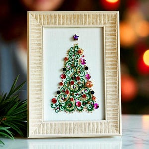 May include: A framed Christmas tree design. The tree is crafted from green thread and decorated with red, pink, and black beads and sequins. A purple star sits at the top. The frame is a light beige colour.