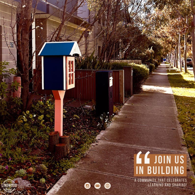 May include: A small, dark blue wooden book exchange box with a peaked roof and a glass-paned door, mounted on a light-colored post supported by three short log segments. The text "JOIN US IN BUILDING A COMMUNITY THAT CELEBRATES LEARNING AND SHARING!" is visible.