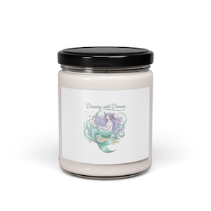 Pode incluir: Um frasco de vela branco com tampa preta. O rótulo apresenta uma ilustração de sereia com cabelo roxo e cauda verde-azulado, rodeada por plantas aquáticas. O texto "Dancing with Dreams" está acima da sereia.