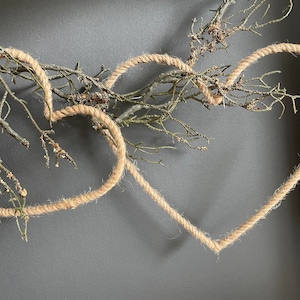 Può includere: Due cuori di corda marrone sono intrecciati con un ramo secco con piccole foglie marroni. I cuori sono appesi a un muro grigio.