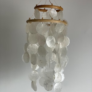 Peut inclure: Un carillon à vent en coquillages blancs avec deux anneaux en bois et une ficelle suspendue au sommet. Les coquillages sont disposés en cascade.