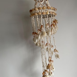 Può includere: Un lampadario decorativo fatto di conchiglie. Il lampadario presenta un design circolare con più fili di conchiglie appese. Le conchiglie sono principalmente di colore crema e marrone chiaro, creando un'estetica naturale e da spiaggia.