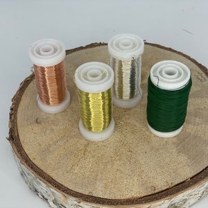 Può includere: Quattro bobine di filo su una superficie di legno. Le bobine sono di colore rame, oro, argento e verde.
