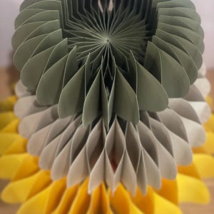 Può includere: Una decorazione in carta a nido d'ape a tre livelli con un design circolare piegato a fisarmonica. Lo strato superiore è verde salvia, lo strato intermedio è beige chiaro e lo strato inferiore è giallo brillante, creando una struttura a ventaglio.