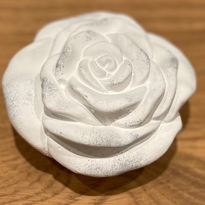 Pode incluir: Uma rosa branca esculpida com pétalas texturizadas. A flor tem uma aparência ligeiramente desgastada, com subtis detalhes cinzentos. A rosa é um objeto decorativo, possivelmente para decoração de casa ou um projeto artesanal.