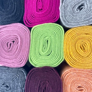 Op de afbeelding: Negen rollen viltstof in verschillende kleuren, waaronder zwart, roze, groen, geel, grijs, bordeauxrood en oranje. De rollen zijn in een rasterpatroon gerangschikt.