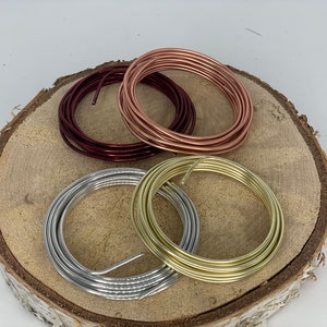 Könnte beinhalten: Vier Spulen Draht in verschiedenen Farben: Silber, Gold, Roségold und Burgunder. Der Draht ist aufgerollt und auf einer Holzoberfläche angeordnet.