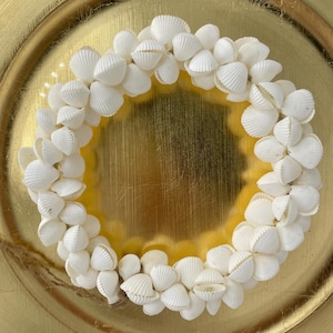 May include: A circular wreath composed of small, white seashells. The wreath is set against a gold-coloured, textured background. A natural twine loop is attached to the wreath.