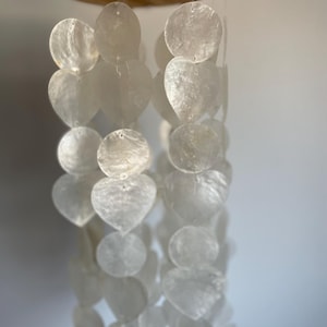 Può includere: Una giostrina sospesa con molteplici fili di dischi di conchiglia bianchi, traslucidi e iridescenti, e pezzi a forma di cuore appesi a un supporto circolare in legno. Sottili filamenti trasparenti sostengono gli elementi naturali.