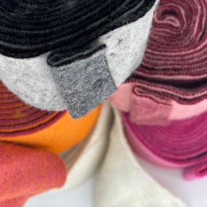 Könnte beinhalten: Nahaufnahme von fünf Rollen Filzstoff in verschiedenen Farben, darunter Schwarz, Grau, Orange, Rosa und Weiß. Die Rollen sind übereinander gestapelt, wobei die schwarze Rolle oben liegt.